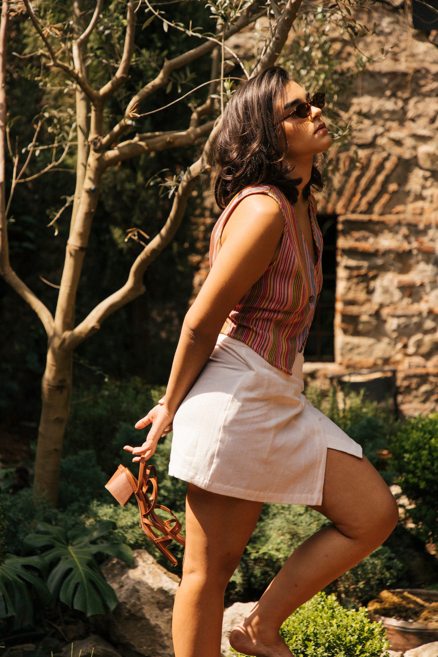Woman in a striped vest and white wrap skirt standing outdoors near a stone building.